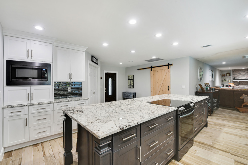 Large contemporary l-shaped eat-in kitchen in Miami with a farmhouse sink, shaker cabinets, grey cabinets, granite benchtops, black splashback, glass tile splashback, stainless steel appliances, ceramic floors, with island, grey floor and multi-coloured benchtop.