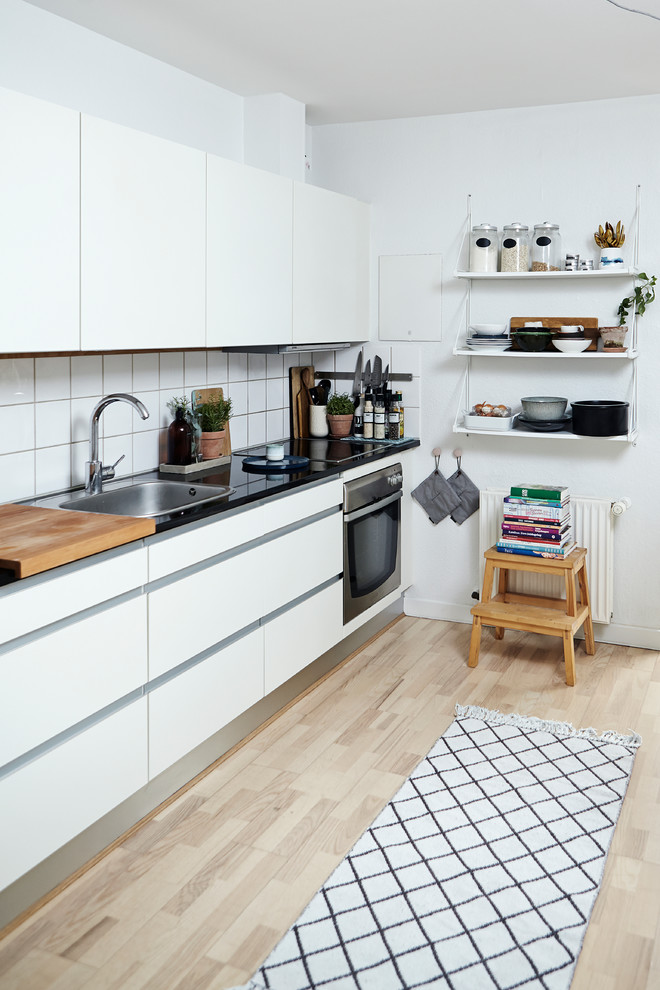 This is an example of a mid-sized scandinavian kitchen in Wiltshire with a drop-in sink, flat-panel cabinets, white cabinets, white splashback, porcelain splashback, light hardwood floors and no island.