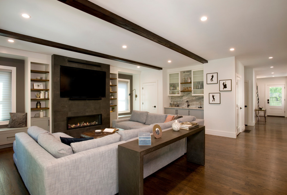 Cette image montre un grand salon traditionnel ouvert avec un mur gris, une cheminée ribbon, un manteau de cheminée en carrelage, un téléviseur encastré, un sol marron et poutres apparentes.