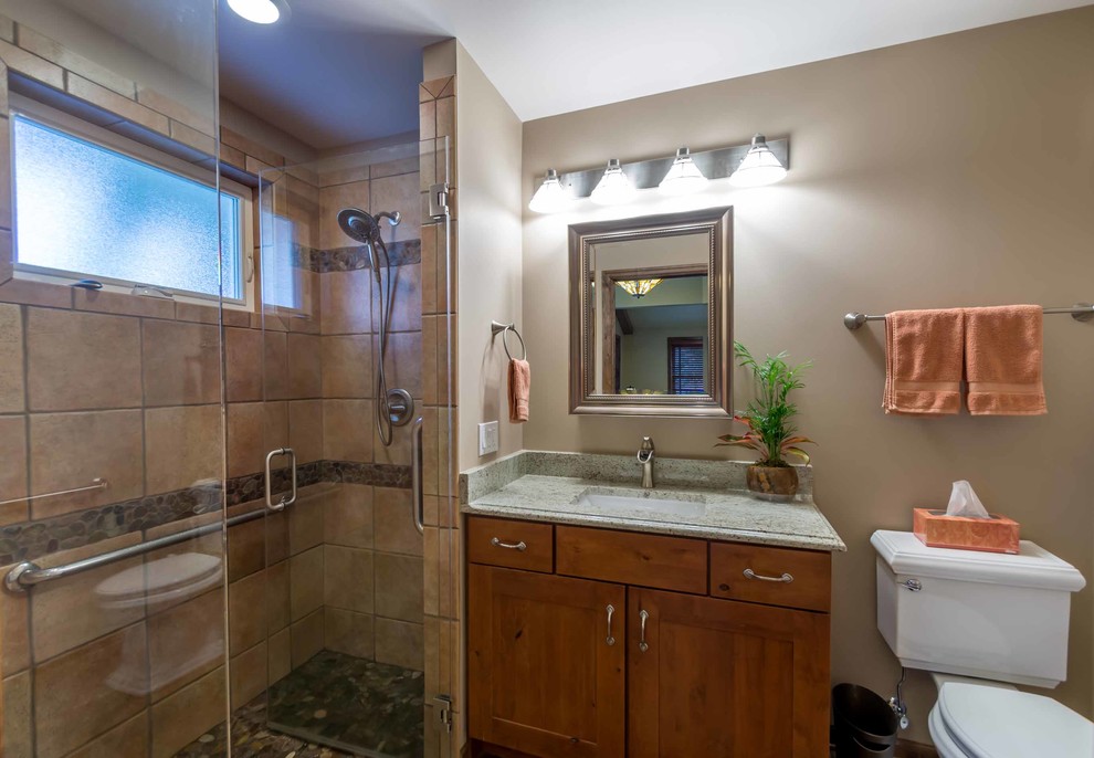 This is an example of a small country 3/4 bathroom in Chicago with shaker cabinets, medium wood cabinets, an alcove shower, a two-piece toilet, brown tile, stone tile, beige walls, ceramic floors, an undermount sink, granite benchtops, a single vanity, a freestanding vanity, grey benchtops, a hinged shower door, brown floor, wallpaper and wallpaper.