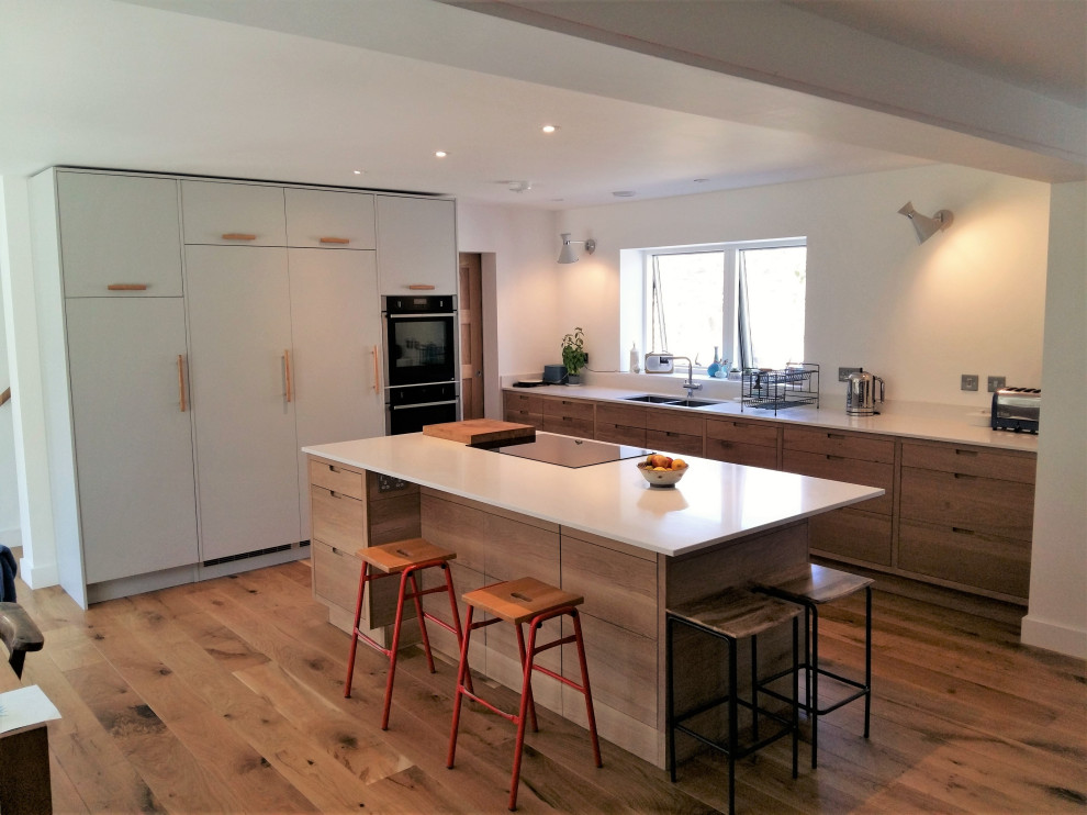 This is an example of a large contemporary l-shaped open plan kitchen in Cornwall with a double-bowl sink, flat-panel cabinets, medium wood cabinets, solid surface benchtops, black appliances, medium hardwood floors, a peninsula, brown floor and white benchtop.