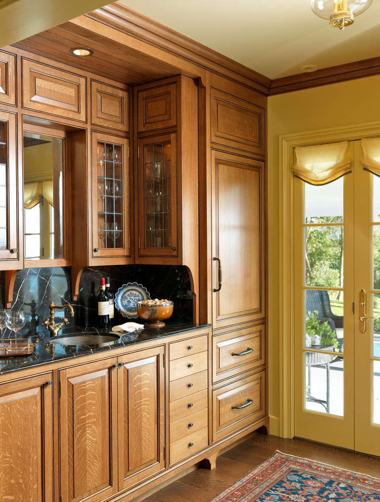 This is an example of a large traditional galley wet bar in Boston with an undermount sink, raised-panel cabinets, medium wood cabinets, marble benchtops, black splashback and dark hardwood floors.