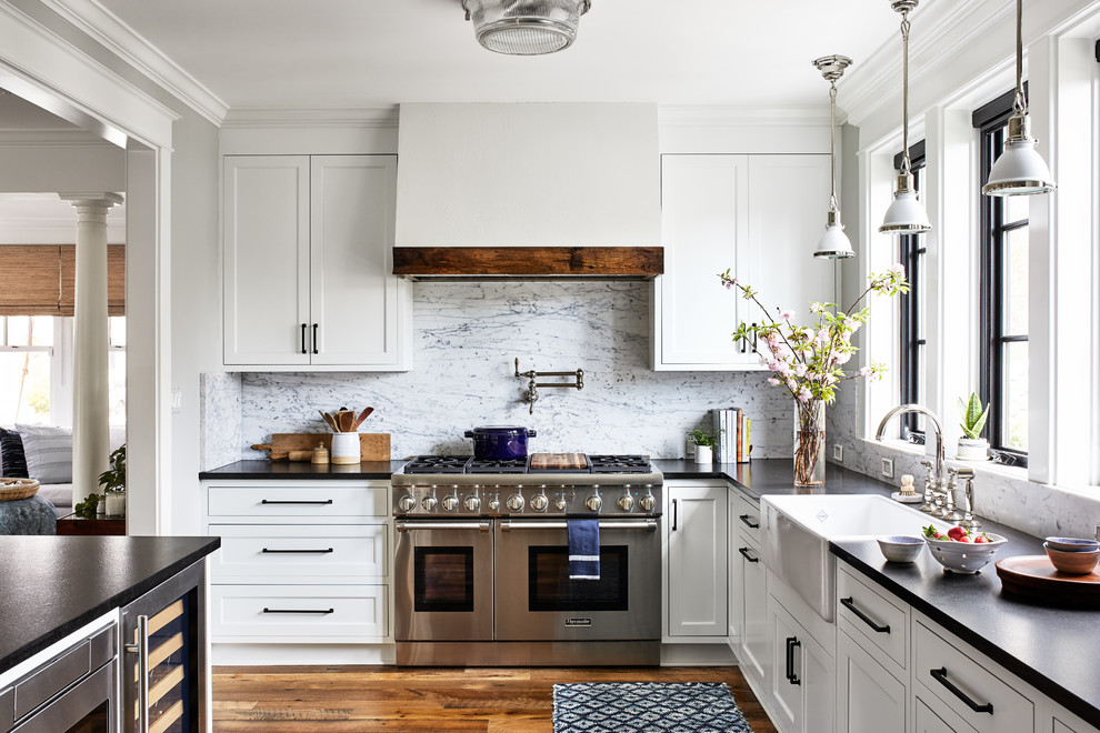 Design ideas for a beach style l-shaped kitchen in DC Metro with a farmhouse sink, shaker cabinets, white cabinets, white splashback, stainless steel appliances, medium hardwood floors, with island, brown floor and black benchtop.