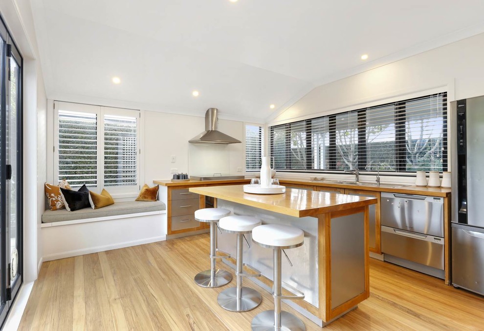 Design ideas for a contemporary l-shaped kitchen in Auckland with a drop-in sink, flat-panel cabinets, stainless steel cabinets, wood benchtops, glass sheet splashback, medium hardwood floors, with island and brown benchtop.