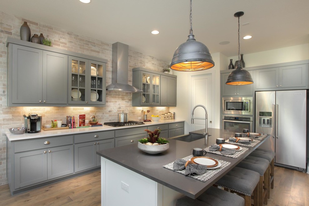 This is an example of a transitional l-shaped kitchen in Austin with a farmhouse sink, shaker cabinets, grey cabinets, beige splashback, stainless steel appliances, medium hardwood floors, with island, brown floor and white benchtop.