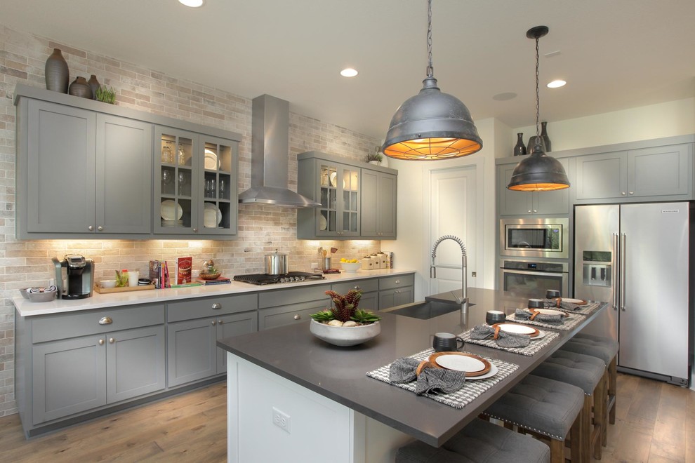 Transitional l-shaped medium tone wood floor and brown floor kitchen photo in Austin with a farmhouse sink, shaker cabinets, gray cabinets, beige backsplash, stainless steel appliances, an island and white countertops