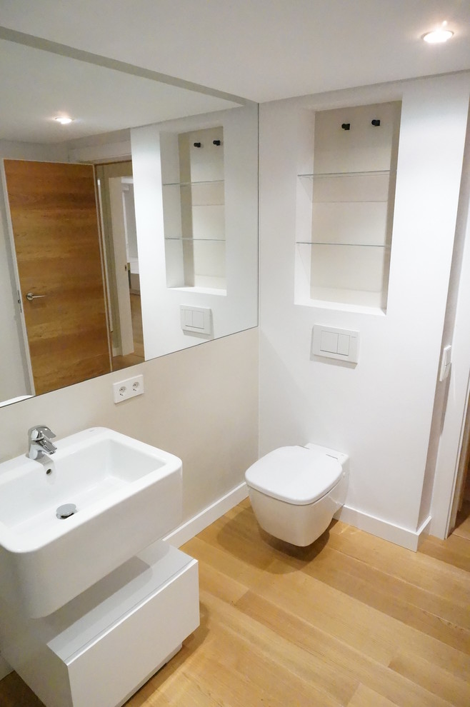 This is an example of a mid-sized contemporary powder room in Valencia with flat-panel cabinets, white cabinets, a wall-mount toilet, white walls, medium hardwood floors and an integrated sink.
