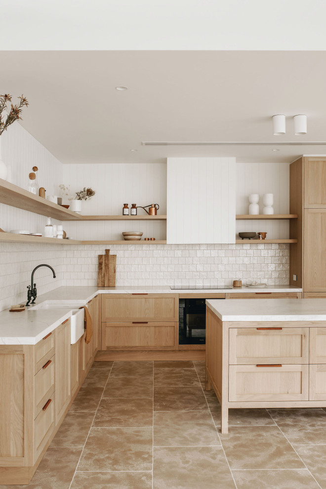 Large scandinavian l-shaped open plan kitchen in Adelaide with a farmhouse sink, recessed-panel cabinets, light wood cabinets, quartz benchtops, beige splashback, ceramic splashback, black appliances, limestone floors, with island and white benchtop.