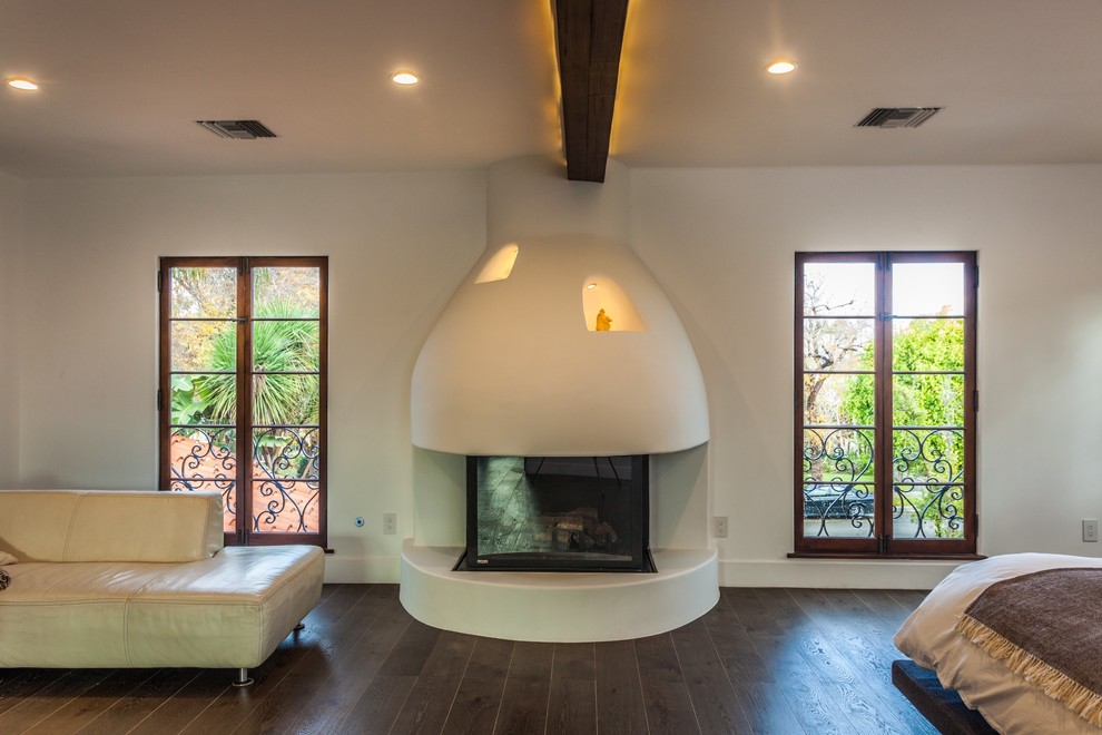 This is an example of a mid-sized mediterranean master bedroom in Los Angeles with white walls, dark hardwood floors, a standard fireplace, a plaster fireplace surround and brown floor.