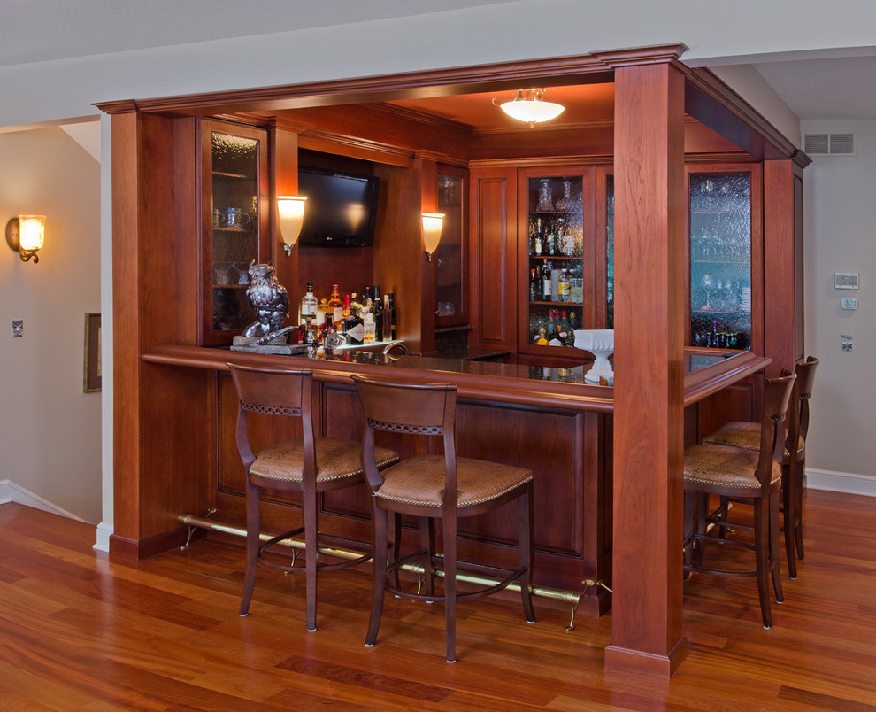This is an example of a mid-sized traditional l-shaped seated home bar in Other with an undermount sink, glass-front cabinets, dark wood cabinets, granite benchtops, brown splashback, timber splashback and medium hardwood floors.