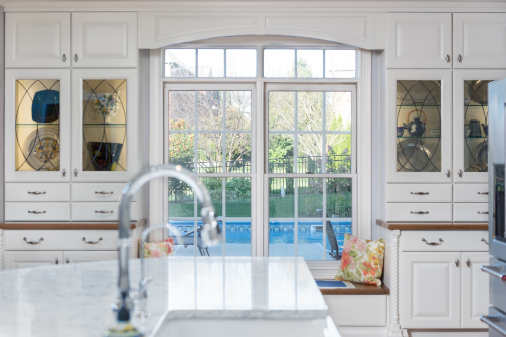 Large transitional single-wall light wood floor and brown floor open concept kitchen photo with a farmhouse sink, raised-panel cabinets, white cabinets, quartz countertops, white backsplash, ceramic backsplash, stainless steel appliances, an island and white countertops