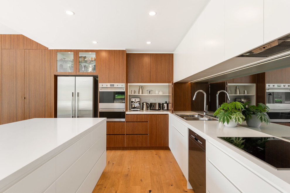 This is an example of a contemporary l-shaped kitchen in Sydney with a double-bowl sink, flat-panel cabinets, medium wood cabinets, mirror splashback, stainless steel appliances, medium hardwood floors, with island, brown floor and white benchtop.