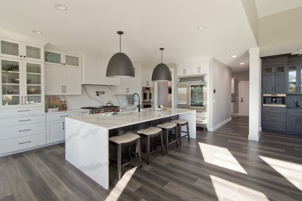 Transitional vinyl floor and brown floor open concept kitchen photo with an undermount sink, recessed-panel cabinets, white cabinets, quartz countertops, white backsplash, stone slab backsplash, stainless steel appliances, an island and white countertops