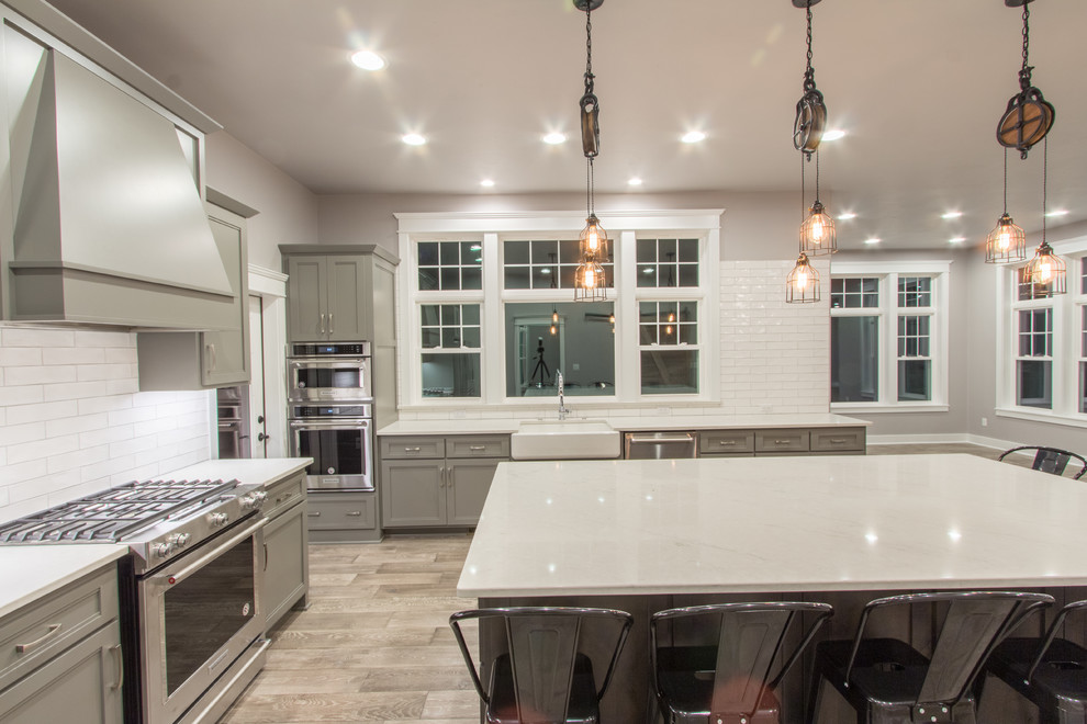 Inspiration for a mid-sized transitional l-shaped open plan kitchen in Milwaukee with a farmhouse sink, shaker cabinets, grey cabinets, quartz benchtops, white splashback, subway tile splashback, stainless steel appliances, medium hardwood floors, with island and grey floor.