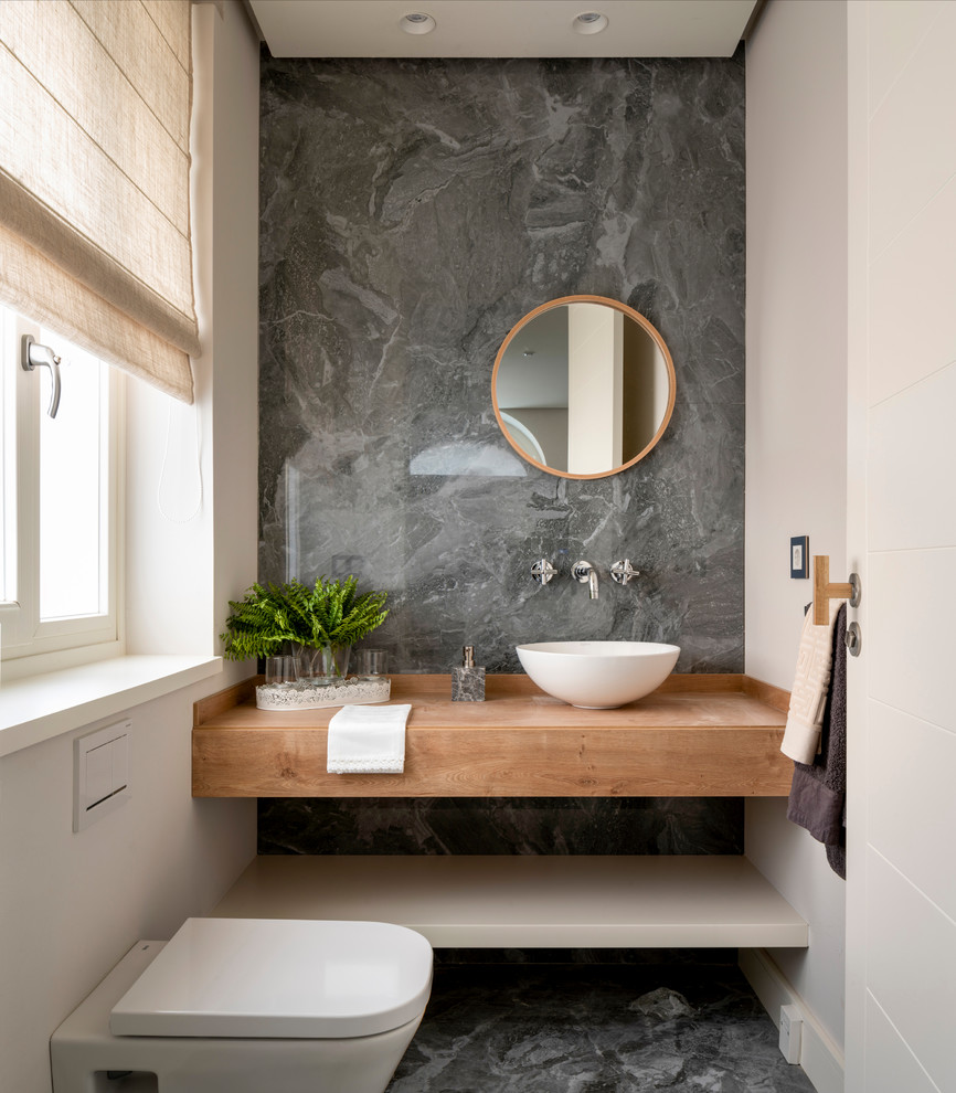 Photo of a mid-sized scandinavian powder room in Bilbao with open cabinets, light wood cabinets, a wall-mount toilet, gray tile, porcelain tile, beige walls, porcelain floors, a vessel sink, wood benchtops, grey floor and brown benchtops.