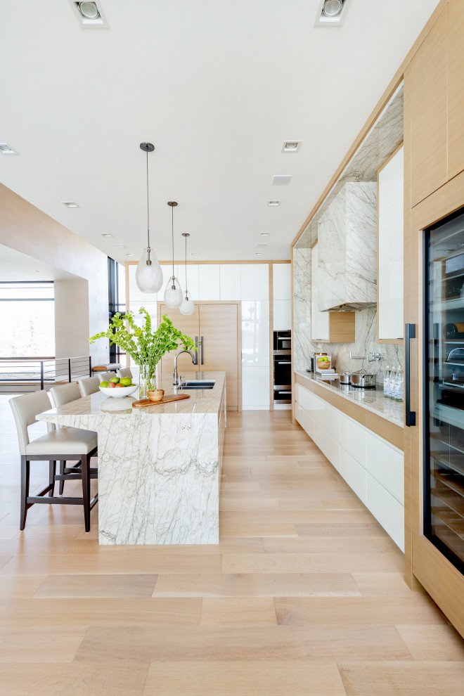 This is an example of a large contemporary l-shaped kitchen pantry in Other with an undermount sink, flat-panel cabinets, light wood cabinets, marble benchtops, white splashback, stone slab splashback, panelled appliances, light hardwood floors, with island, beige floor and white benchtop.