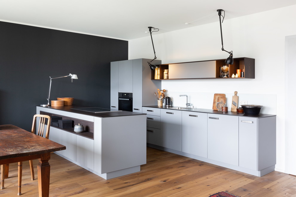 Mid-sized contemporary galley eat-in kitchen in Cologne with a single-bowl sink, flat-panel cabinets, grey cabinets, white splashback, black appliances, light hardwood floors, a peninsula and black benchtop.