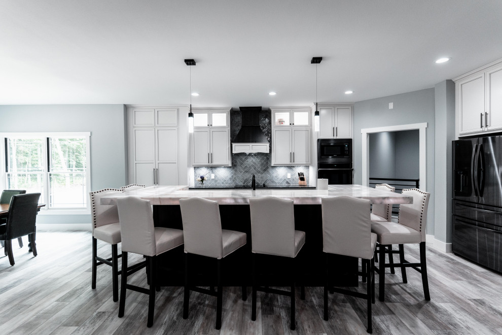 Example of a huge transitional l-shaped laminate floor, gray floor and tray ceiling open concept kitchen design with an undermount sink, flat-panel cabinets, white cabinets, quartz countertops, gray backsplash, ceramic backsplash, black appliances and an island