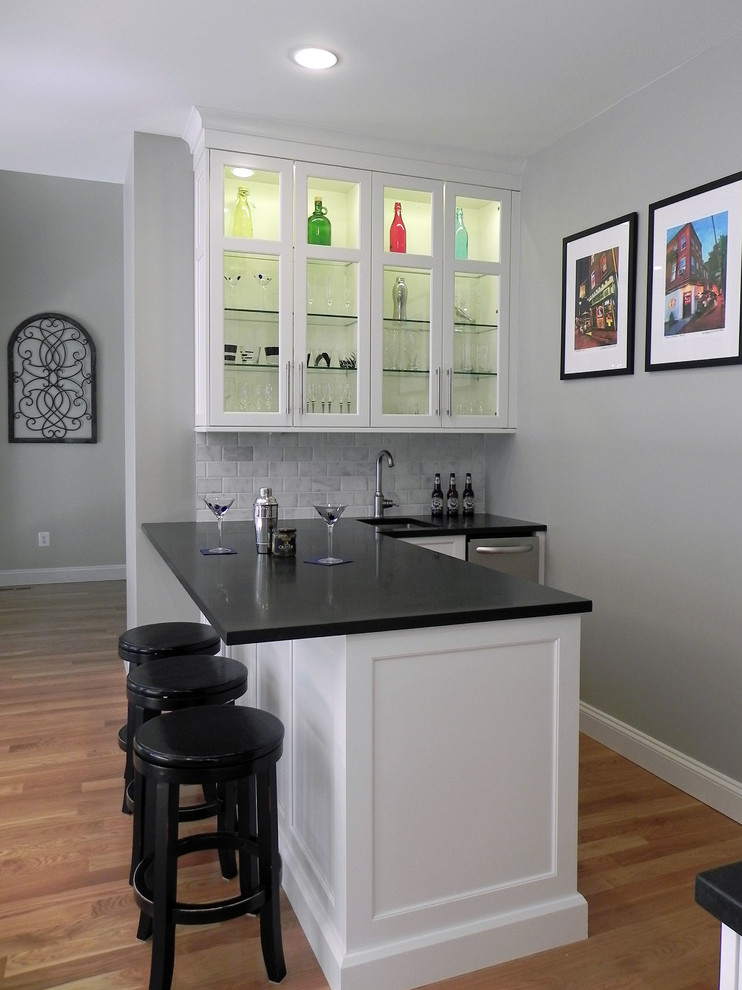 Design ideas for a mid-sized transitional l-shaped wet bar in Baltimore with an undermount sink, shaker cabinets, white cabinets, quartz benchtops, grey splashback, stone tile splashback, medium hardwood floors and brown floor.