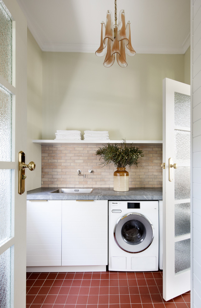 This is an example of a laundry room in Sydney with a single-bowl sink, soapstone benchtops, terra-cotta splashback, green walls, terra-cotta floors and grey benchtop.