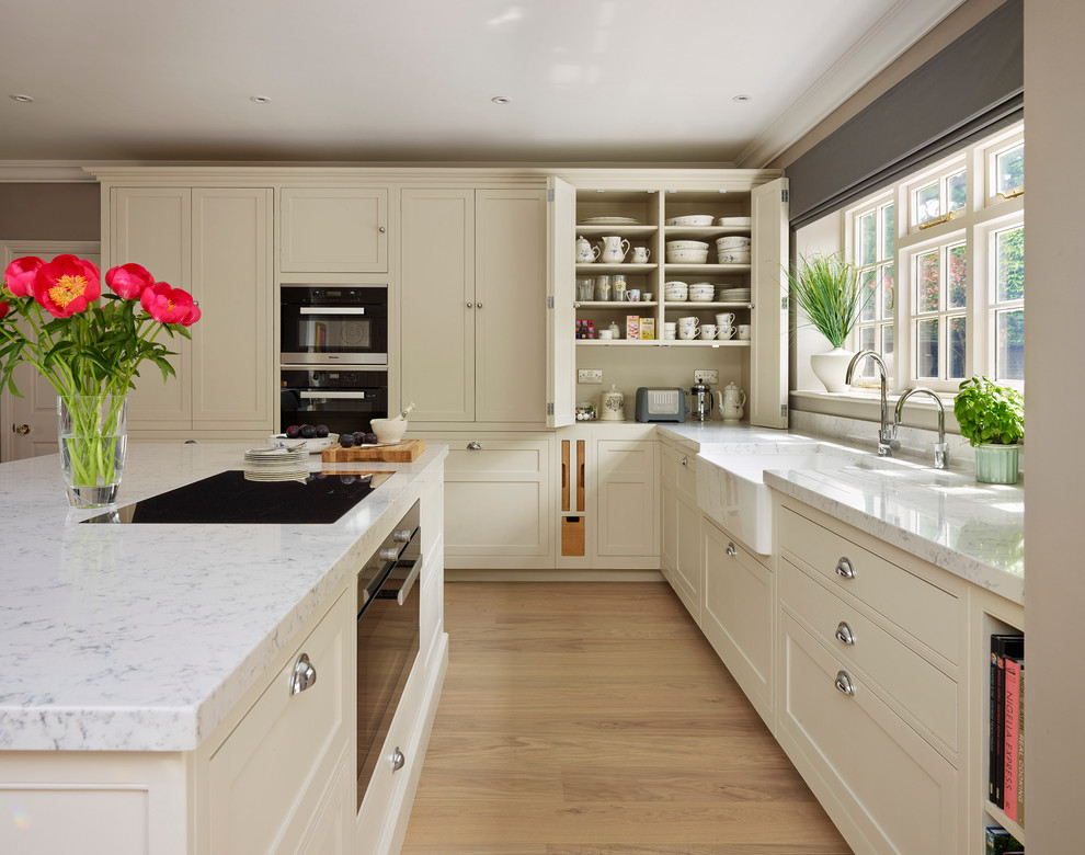 Design ideas for a transitional kitchen in Oxfordshire with a farmhouse sink, flat-panel cabinets, beige cabinets, black appliances, light hardwood floors and with island.