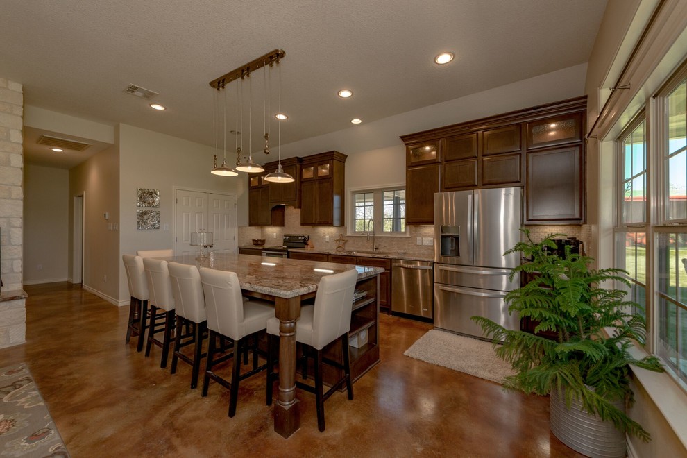 Mid-sized country u-shaped open plan kitchen in Austin with an undermount sink, shaker cabinets, medium wood cabinets, granite benchtops, beige splashback, travertine splashback, stainless steel appliances, concrete floors, with island and brown floor.