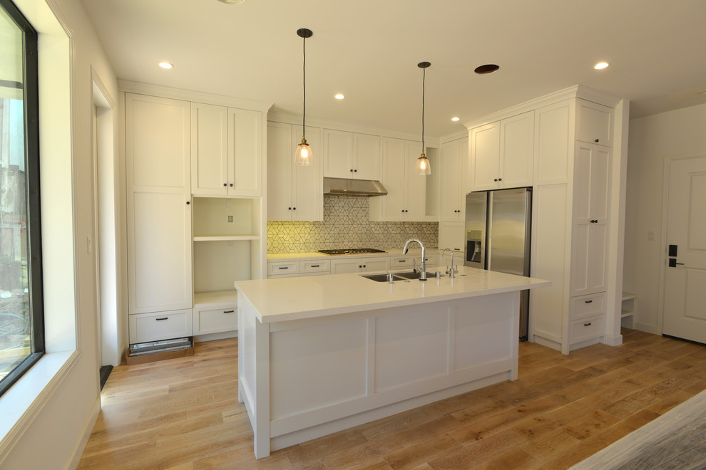 Photo of a transitional l-shaped open plan kitchen in Los Angeles with a double-bowl sink, shaker cabinets, white cabinets, quartzite benchtops, white splashback, ceramic splashback, stainless steel appliances, light hardwood floors and with island.