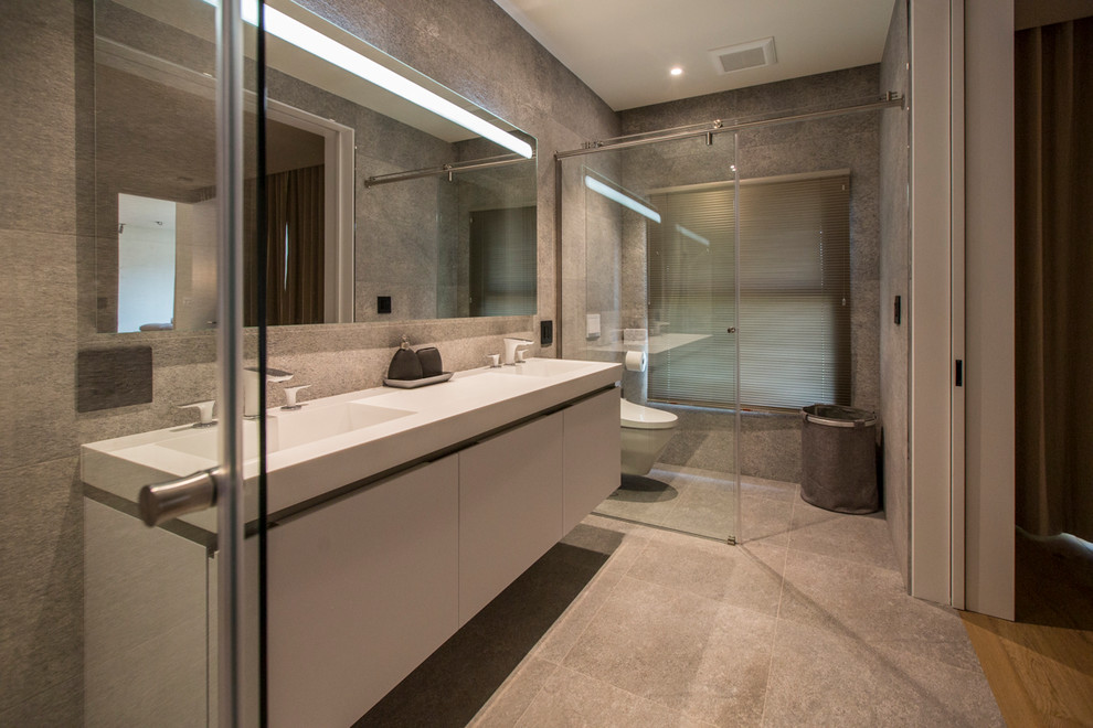 This is an example of a mid-sized contemporary master bathroom in San Francisco with flat-panel cabinets, white cabinets, a curbless shower, a wall-mount toilet, gray tile, cement tile, grey walls, concrete floors, an integrated sink, solid surface benchtops, grey floor, a sliding shower screen and white benchtops.