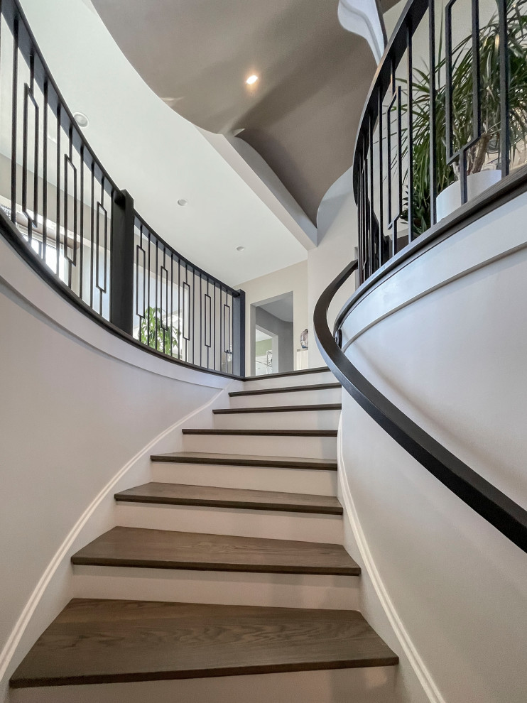 Стильный дизайн: огромная изогнутая лестница в стиле неоклассика (современная классика) с деревянными ступенями, крашенными деревянными подступенками, перилами из смешанных материалов и обоями на стенах - последний тренд
