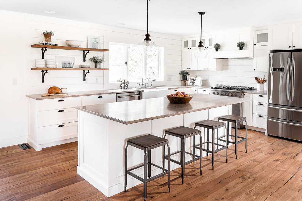 Photo of a large country l-shaped eat-in kitchen in Other with a farmhouse sink, shaker cabinets, white cabinets, marble benchtops, white splashback, stainless steel appliances, medium hardwood floors, with island, brown floor and grey benchtop.