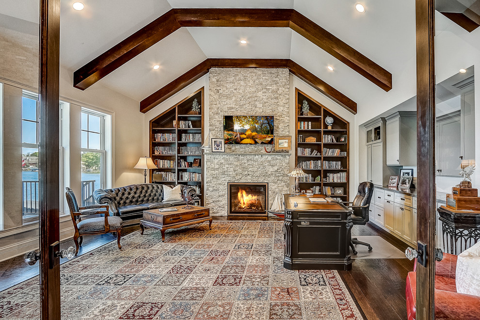 This is an example of an expansive beach style home office in Milwaukee with a library, white walls, dark hardwood floors, a standard fireplace, a stone fireplace surround, a freestanding desk and brown floor.