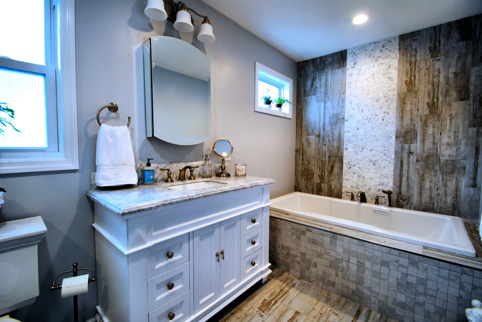 This is an example of a mid-sized country master bathroom in New York with furniture-like cabinets, white cabinets, a drop-in tub, an alcove shower, a two-piece toilet, brown tile, pebble tile, grey walls, porcelain floors, an undermount sink, marble benchtops, brown floor, a sliding shower screen and multi-coloured benchtops.