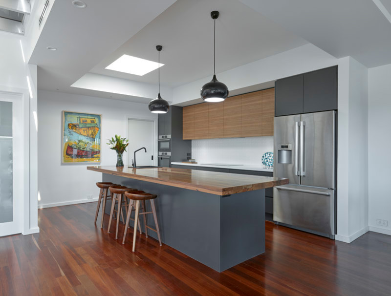 Photo of a large eclectic galley open plan kitchen in Other with a single-bowl sink, flat-panel cabinets, grey cabinets, wood benchtops, white splashback, porcelain splashback, stainless steel appliances, medium hardwood floors, with island and brown benchtop.