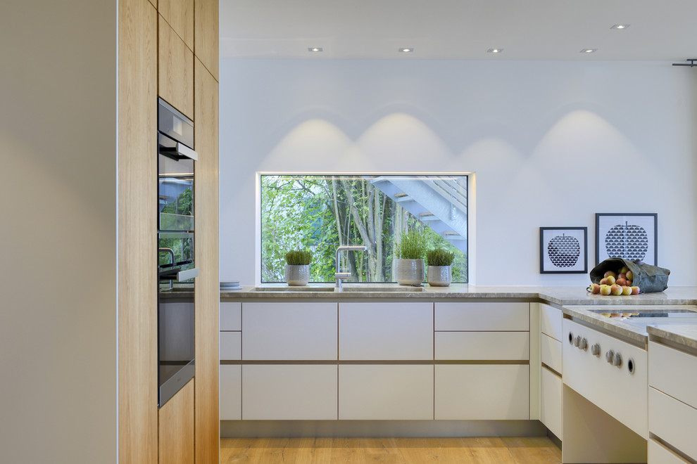 Large contemporary l-shaped open plan kitchen in Other with an integrated sink, flat-panel cabinets, white cabinets, limestone benchtops, black appliances, medium hardwood floors and white splashback.