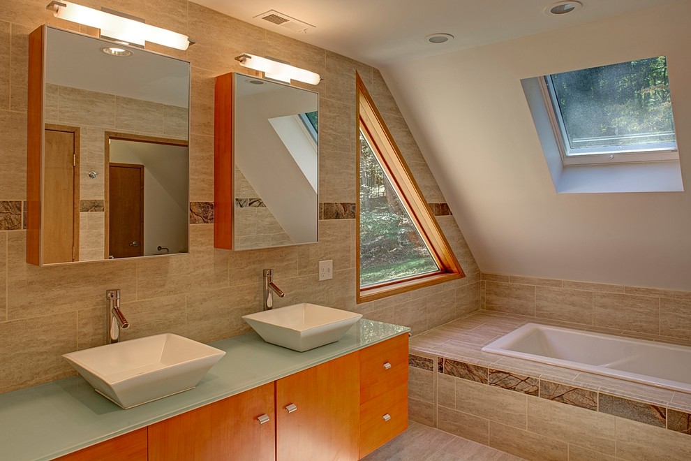 This is an example of a mid-sized modern master bathroom in Baltimore with flat-panel cabinets, medium wood cabinets, a drop-in tub, beige tile, porcelain tile, beige walls, limestone floors, a vessel sink and glass benchtops.