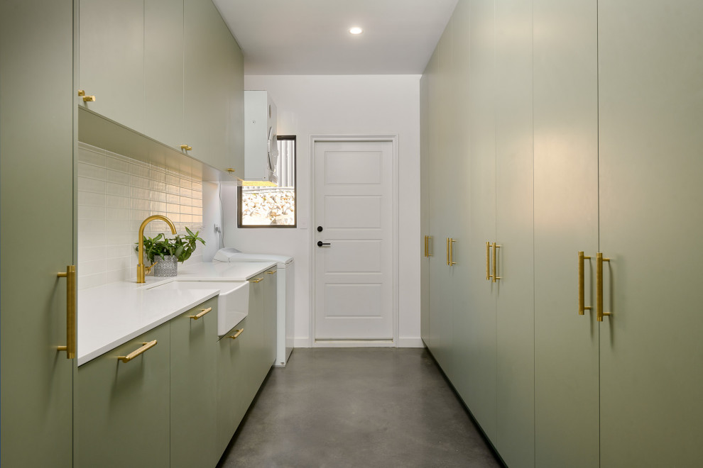 Design ideas for a large classic galley separated utility room in Perth with a belfast sink, flat-panel cabinets, green cabinets, engineered stone countertops, white splashback, metro tiled splashback, white walls, concrete flooring, a stacked washer and dryer, grey floors, white worktops and brick walls.