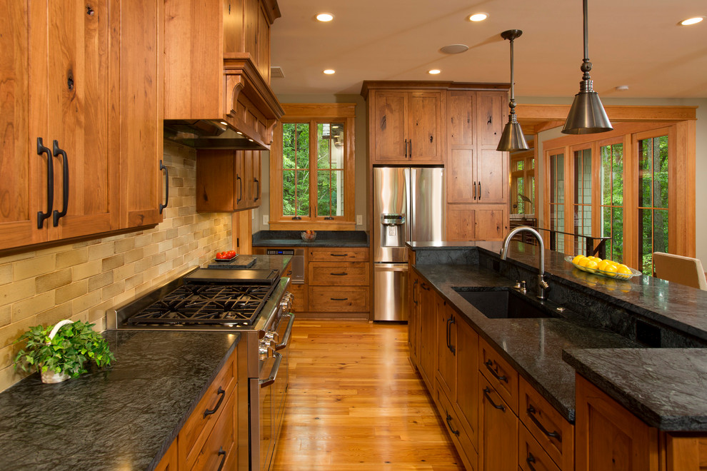 Inspiration for a large country l-shaped eat-in kitchen in DC Metro with an undermount sink, shaker cabinets, medium wood cabinets, beige splashback, stainless steel appliances, medium hardwood floors, with island, soapstone benchtops, subway tile splashback and brown floor.