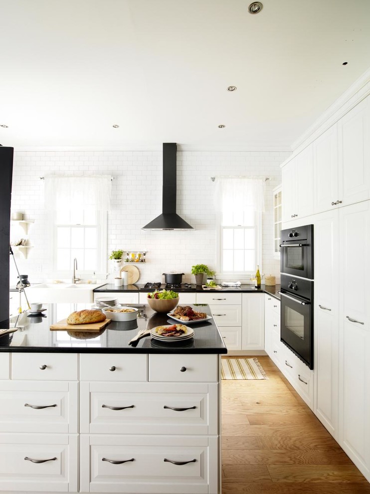 This is an example of a large contemporary u-shaped eat-in kitchen in Grand Rapids with a farmhouse sink, raised-panel cabinets, white cabinets, quartz benchtops, white splashback, subway tile splashback, black appliances, light hardwood floors, with island and yellow floor.