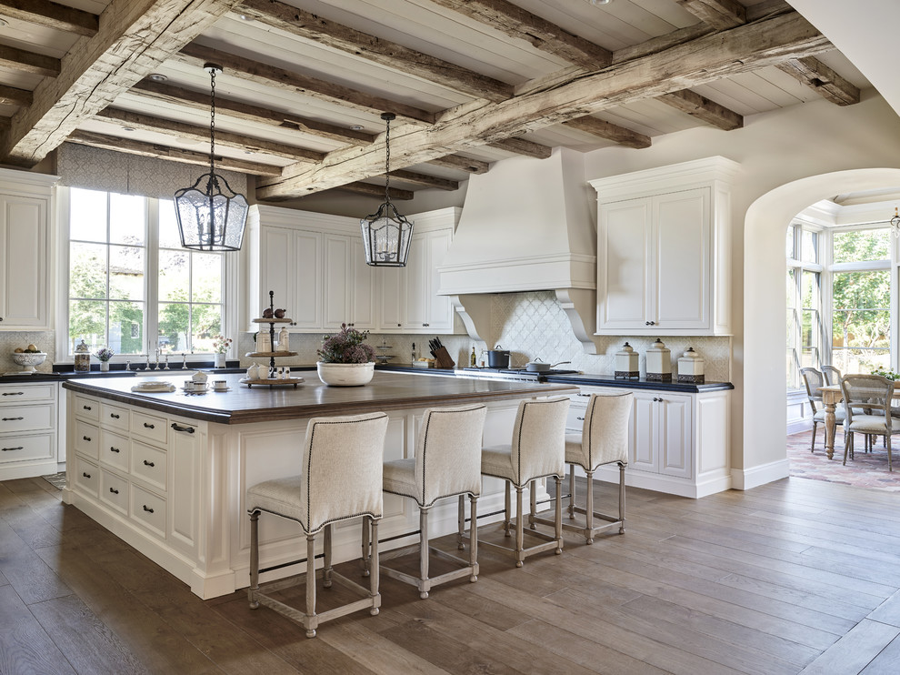 Photo of a large transitional l-shaped kitchen in Phoenix with raised-panel cabinets, beige splashback, with island, white cabinets, a farmhouse sink, soapstone benchtops, ceramic splashback, medium hardwood floors and brown floor.