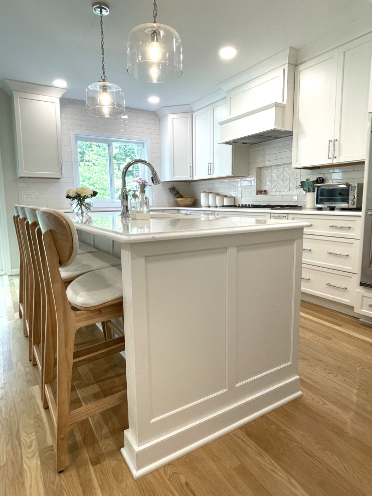 Inspiration for a large transitional l-shaped eat-in kitchen in Detroit with an undermount sink, shaker cabinets, white cabinets, quartz benchtops, white splashback, ceramic splashback, stainless steel appliances, medium hardwood floors, with island, brown floor and white benchtop.