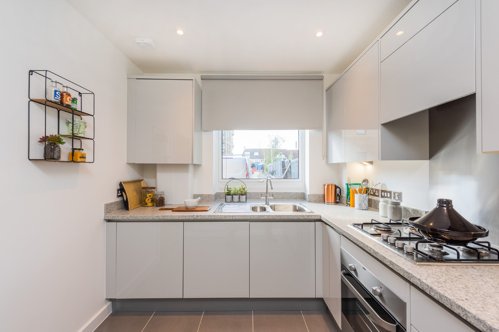This is an example of a contemporary l-shaped separate kitchen in London with a drop-in sink, flat-panel cabinets, white cabinets, granite benchtops, metallic splashback, metal splashback, stainless steel appliances, no island, grey floor and beige benchtop.