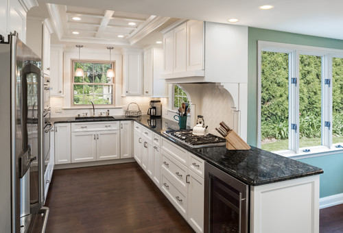 Bright white kitchen cabinet set with shaker doors, black granite countertops, a gas cooktop, coffered ceiling, and large garden-view windows.