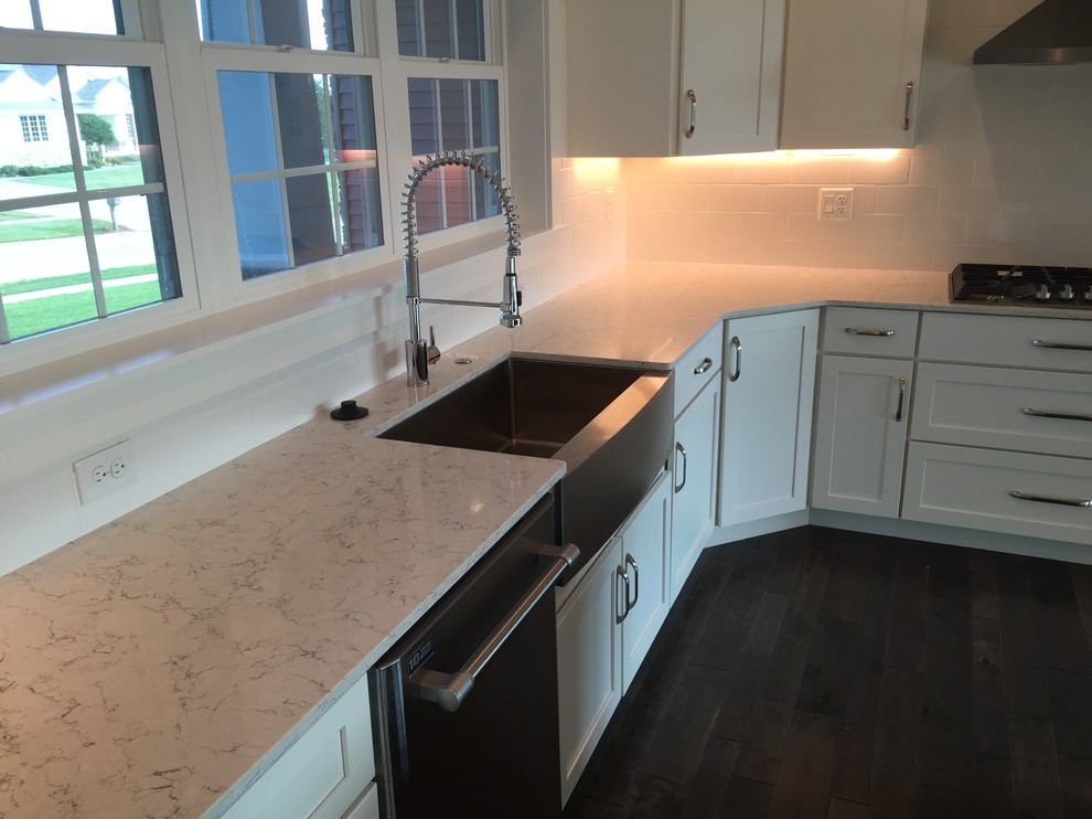 Example of a mid-sized transitional l-shaped dark wood floor and brown floor kitchen design with a farmhouse sink, shaker cabinets, white cabinets, quartz countertops, white backsplash, ceramic backsplash, stainless steel appliances, an island and white countertops