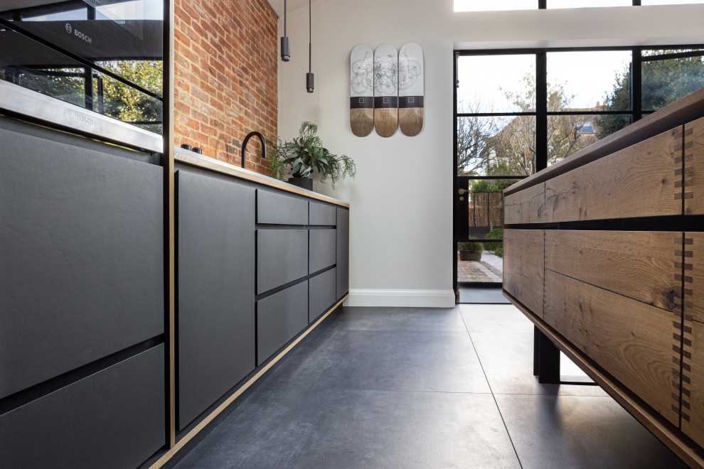 Large contemporary single-wall open plan kitchen in Sussex with a drop-in sink, flat-panel cabinets, distressed cabinets, quartzite benchtops, multi-coloured splashback, brick splashback, black appliances, ceramic floors, with island, grey floor, grey benchtop and vaulted.