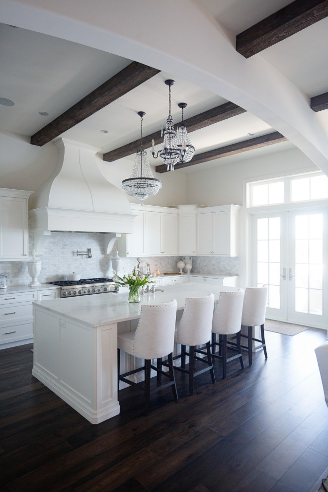 This is an example of a large beach style u-shaped eat-in kitchen in Miami with a farmhouse sink, shaker cabinets, white cabinets, quartzite benchtops, white splashback, ceramic splashback, panelled appliances, dark hardwood floors, with island, brown floor, white benchtop and exposed beam.