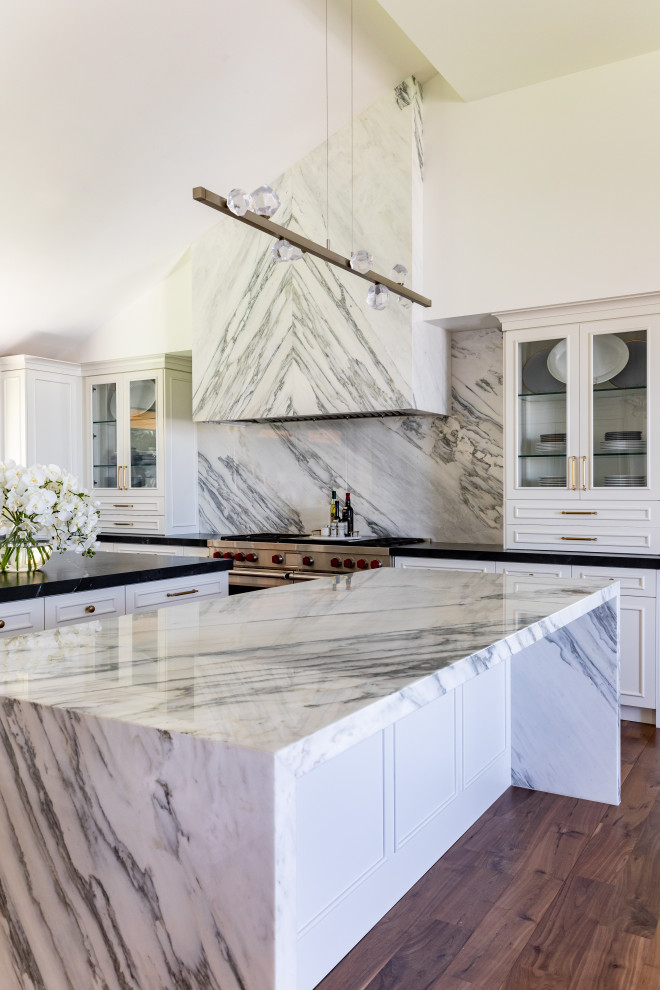 Modern inredning av ett mycket stort vit vitt u-kök, med en undermonterad diskho, släta luckor, vita skåp, marmorbänkskiva, vitt stänkskydd, stänkskydd i marmor, integrerade vitvaror, mellanmörkt trägolv, flera köksöar och brunt golv