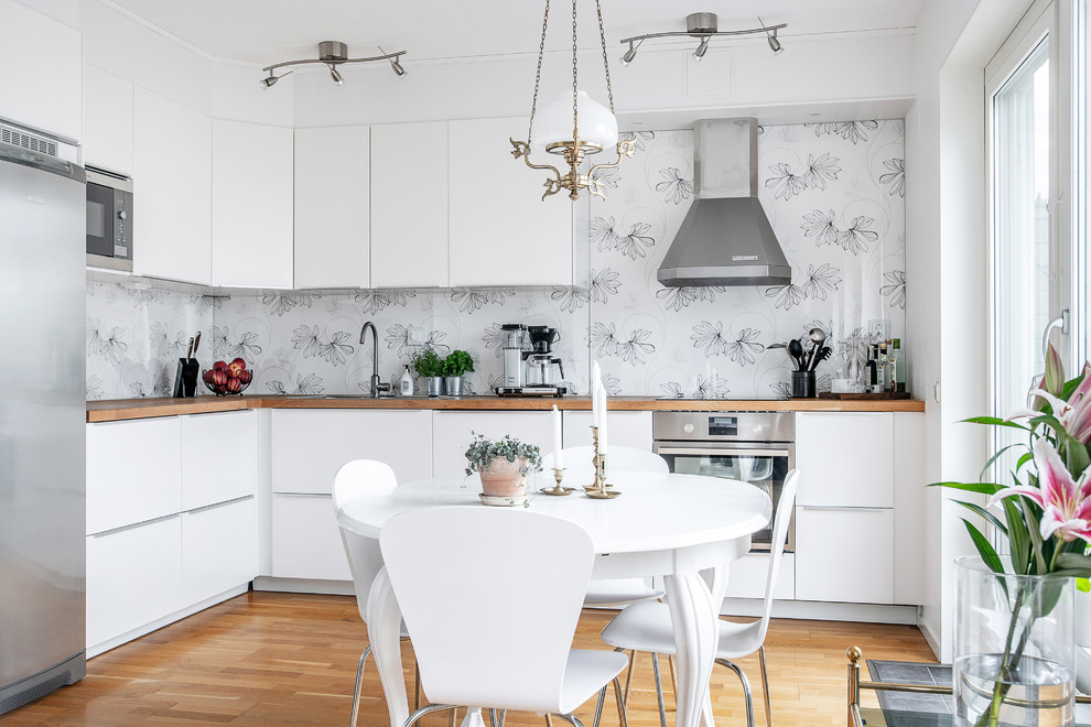 This is an example of a small scandinavian l-shaped eat-in kitchen in Stockholm with flat-panel cabinets, white cabinets, wood benchtops, stainless steel appliances, medium hardwood floors, no island, brown floor and brown benchtop.