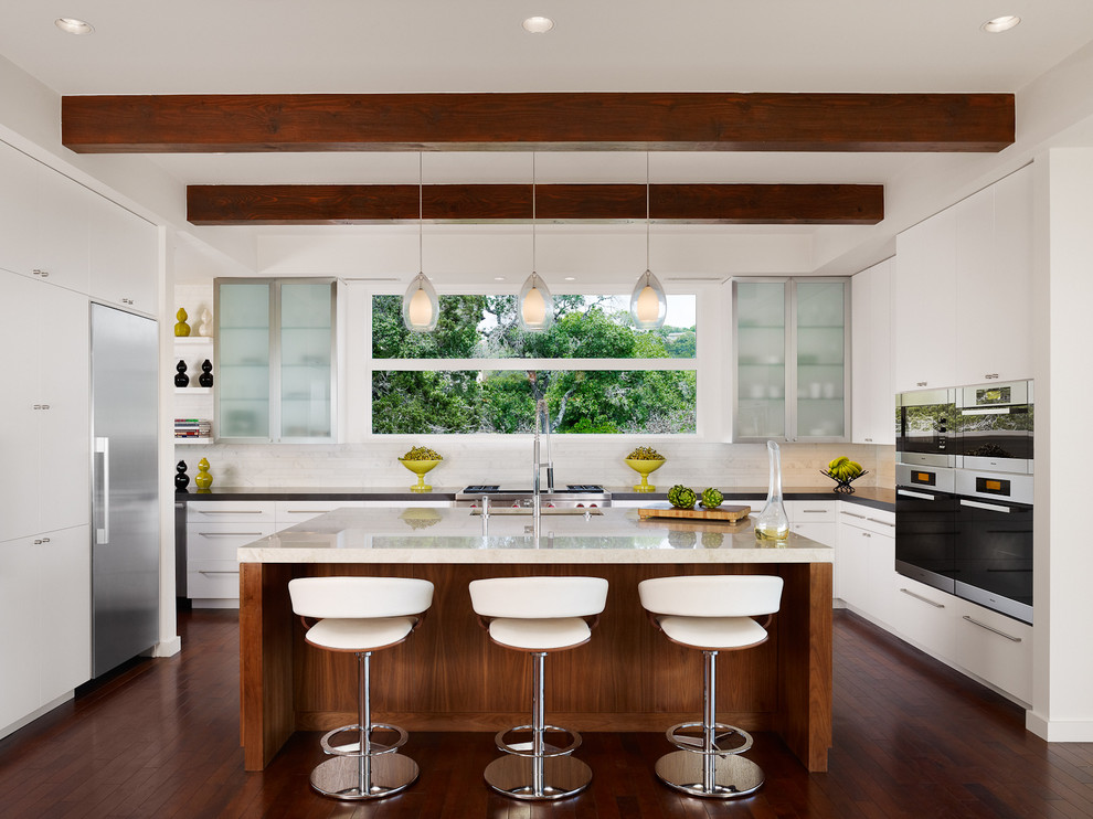 Design ideas for a large contemporary galley open plan kitchen in Austin with an integrated sink, flat-panel cabinets, white cabinets, white splashback, stone tile splashback, stainless steel appliances, dark hardwood floors, with island, marble benchtops and brown floor.