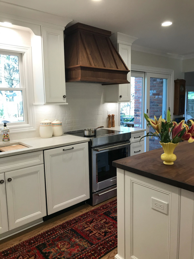 Photo of a large classic galley kitchen/diner in Other with an island, a submerged sink, recessed-panel cabinets, white cabinets, engineered stone countertops, white splashback, brick splashback, stainless steel appliances, medium hardwood flooring, brown floors and white worktops.