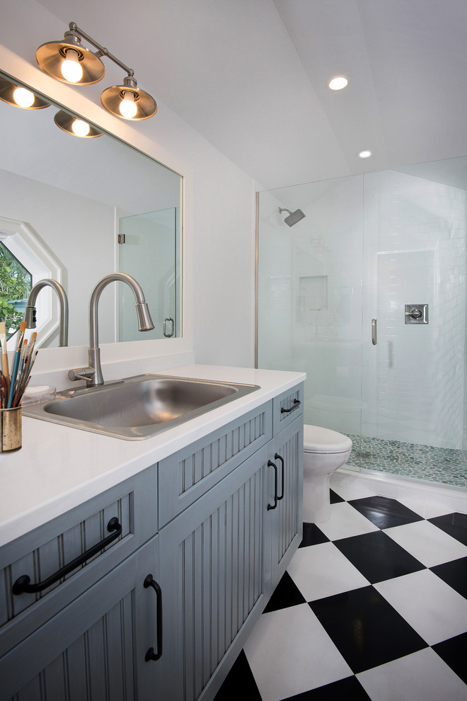 This is an example of a mid-sized beach style 3/4 bathroom in Tampa with beaded inset cabinets, blue cabinets, a shower/bathtub combo, a one-piece toilet, white tile, ceramic tile, white walls, linoleum floors, a drop-in sink, engineered quartz benchtops, white floor, a hinged shower door and white benchtops.
